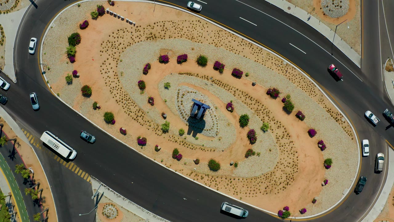 Top-down Los Cabos, Mexico frame showing spaced cars on dark curved lanes beside gravel beds, low shrubs, and a central blue-roofed structure, no visible people.