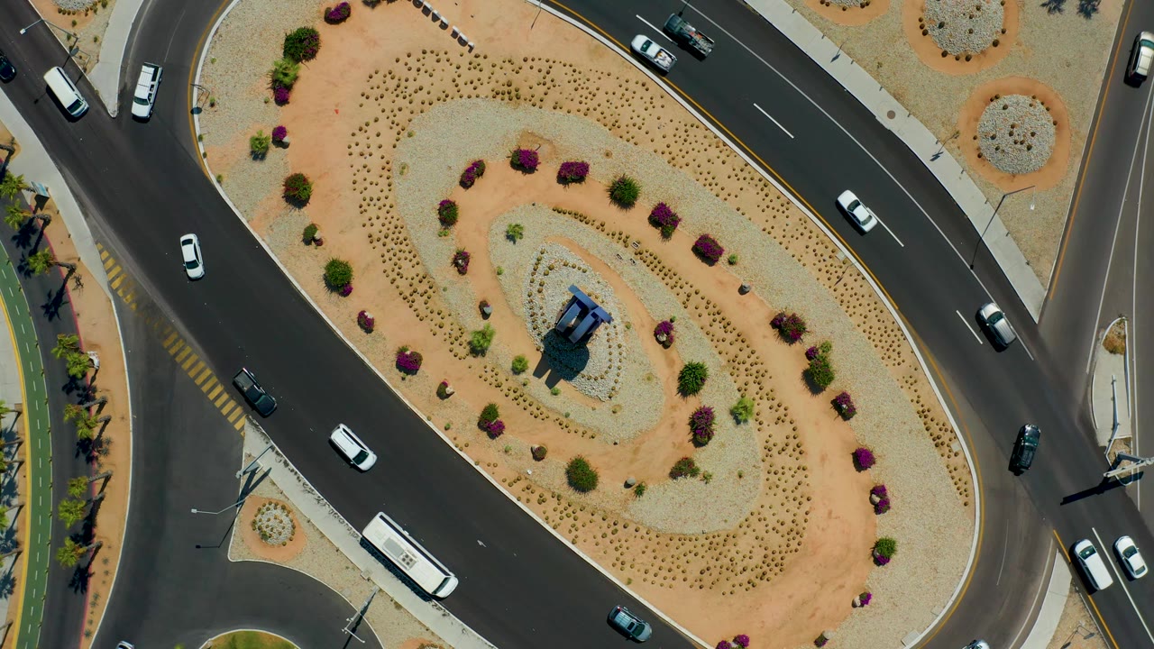 Overhead Los Cabos, Mexico frame showing a white bus, several cars, dark asphalt, pale sidewalks, and a desert-planted traffic island with no visible people.