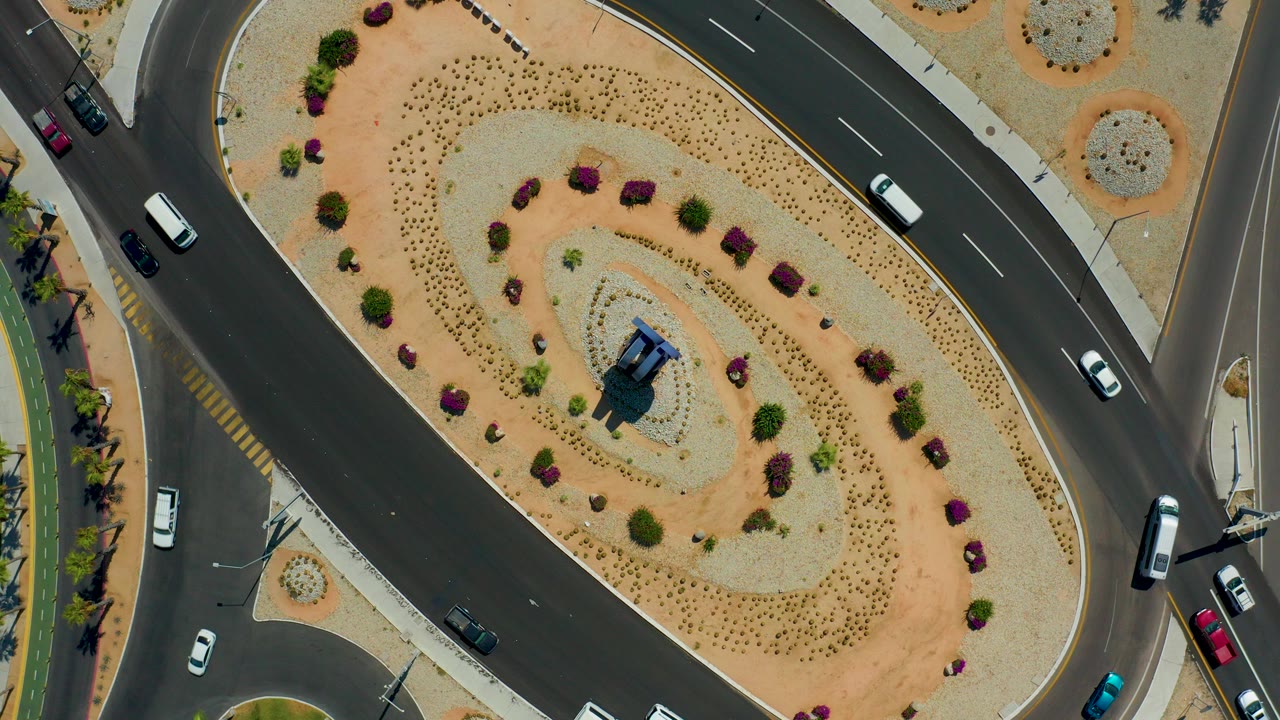 Overhead frame of Los Cabos highway traffic curving around a tan landscaped median with shrubs, palms along the edge, several cars, and no visible people.