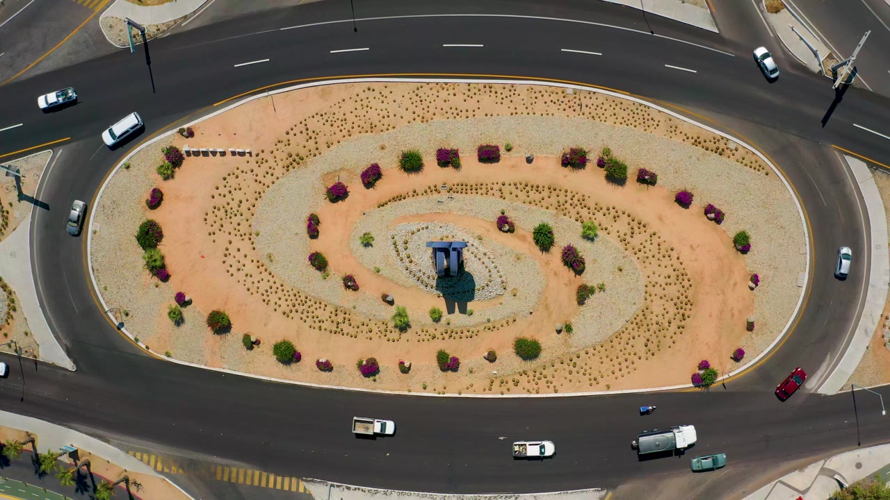 Aerial view in Los Cabos, Mexico, with white cars, a red vehicle, and a larger dark vehicle spaced around the oval road, with no visible people.