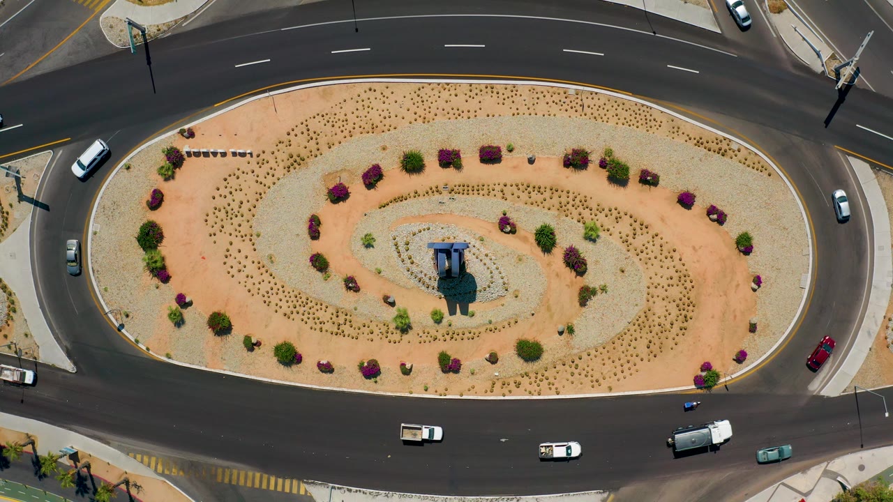 Direct overhead view of vehicles moving around a pale desert-landscaped oval in Los Cabos, Mexico, with the road wrapping the frame and no visible people.