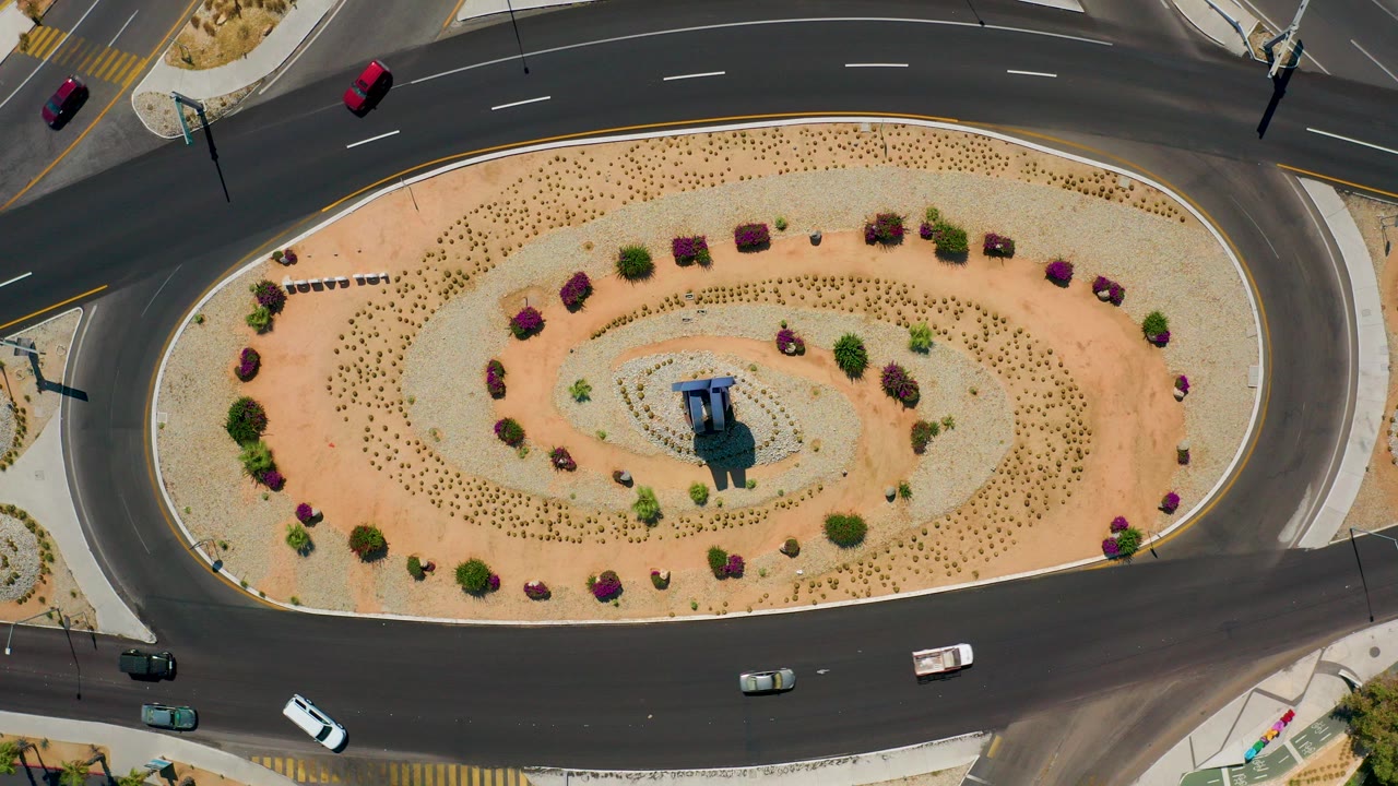 Top-down aerial frame in Los Cabos, Mexico showing cars on dark asphalt around an oval landscaped traffic circle with shrubs, small trees, and no visible people.