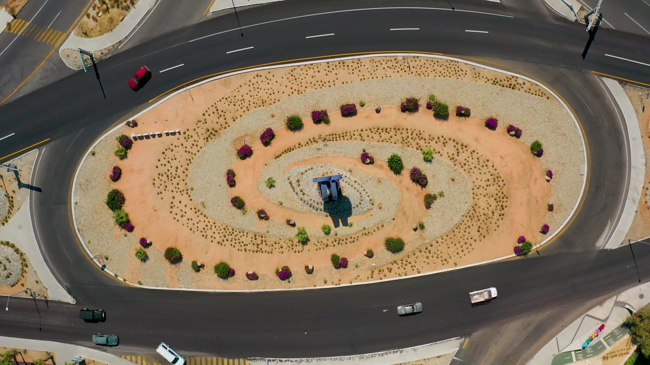 High overhead frame in Los Cabos, Mexico with cars on the curved black roadway and a pale landscaped oval filled with dotted plantings, bushes, and no visible people.