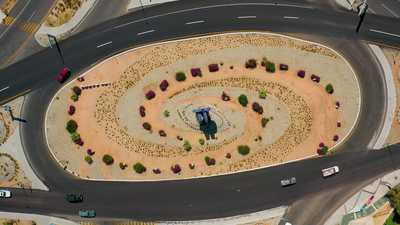 Aerial road frame showing a red car at the left edge, white and green vehicles along the lower lanes, and an oval Los Cabos traffic circle with dry landscaping and no visible people.