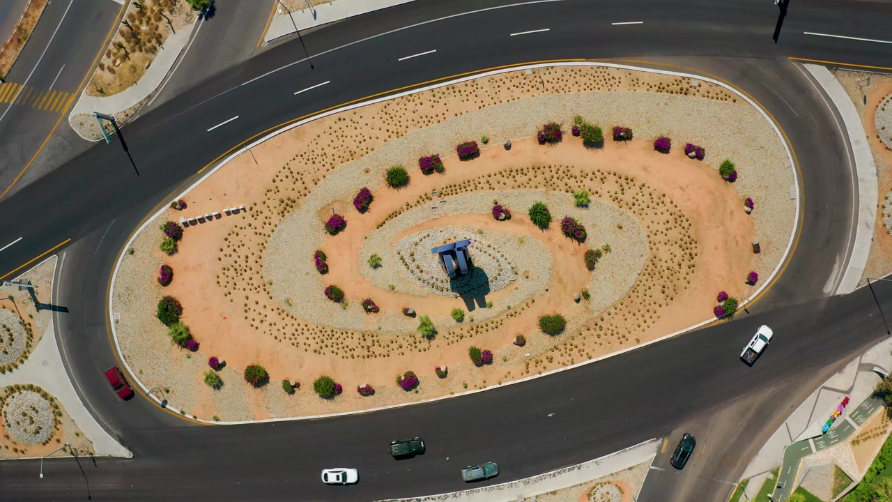 Overhead Los Cabos road view with a white vehicle on the right curve, several cars along the lower road, sandy planting beds, and no visible people.