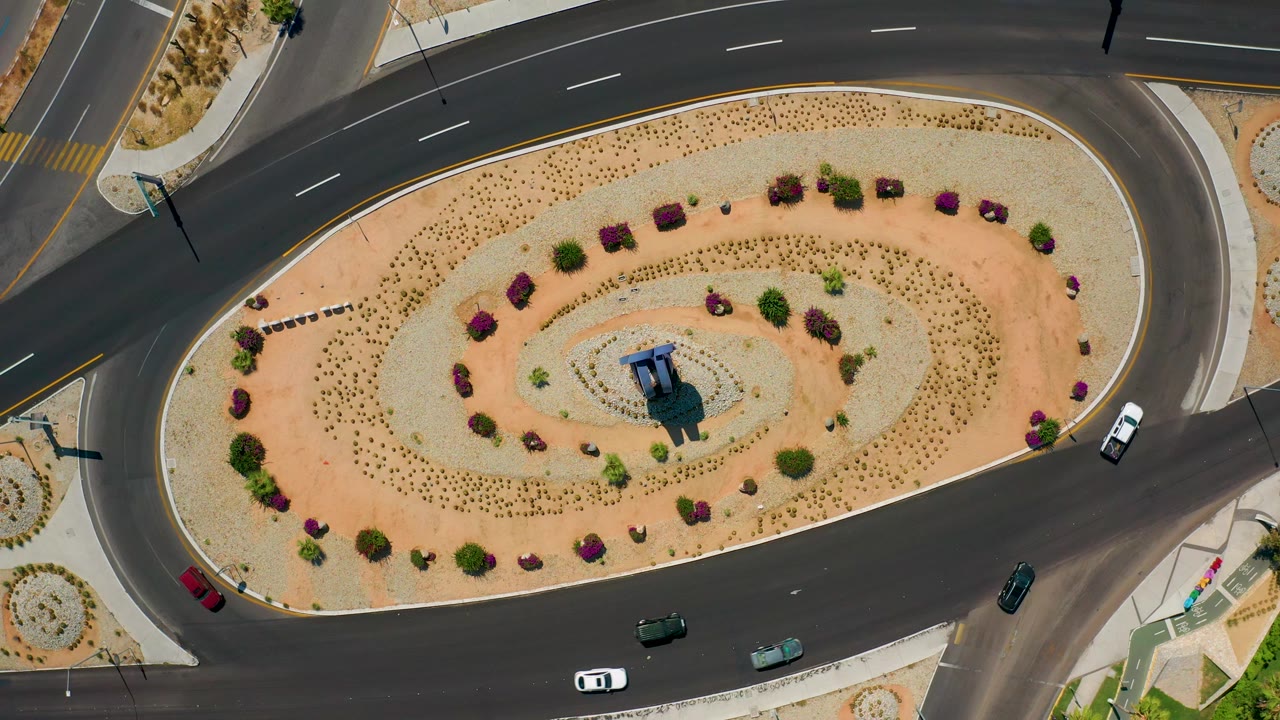 Aerial frame from Los Cabos, Mexico showing a white car near the right edge, multiple vehicles on the lower road, desert plantings, sidewalks, and no visible people.