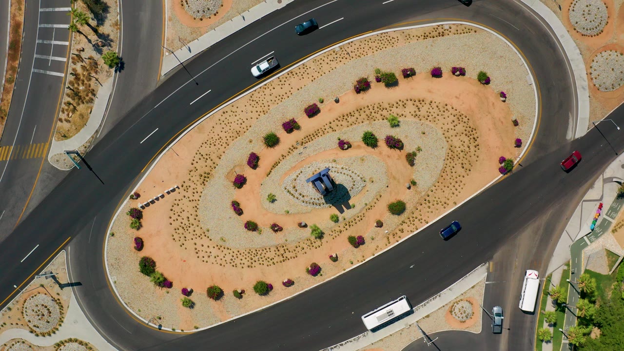 Overhead aerial frame of a Los Cabos, Mexico traffic roundabout with desert landscaping, several cars, a white bus near the lower road, and no visible people.