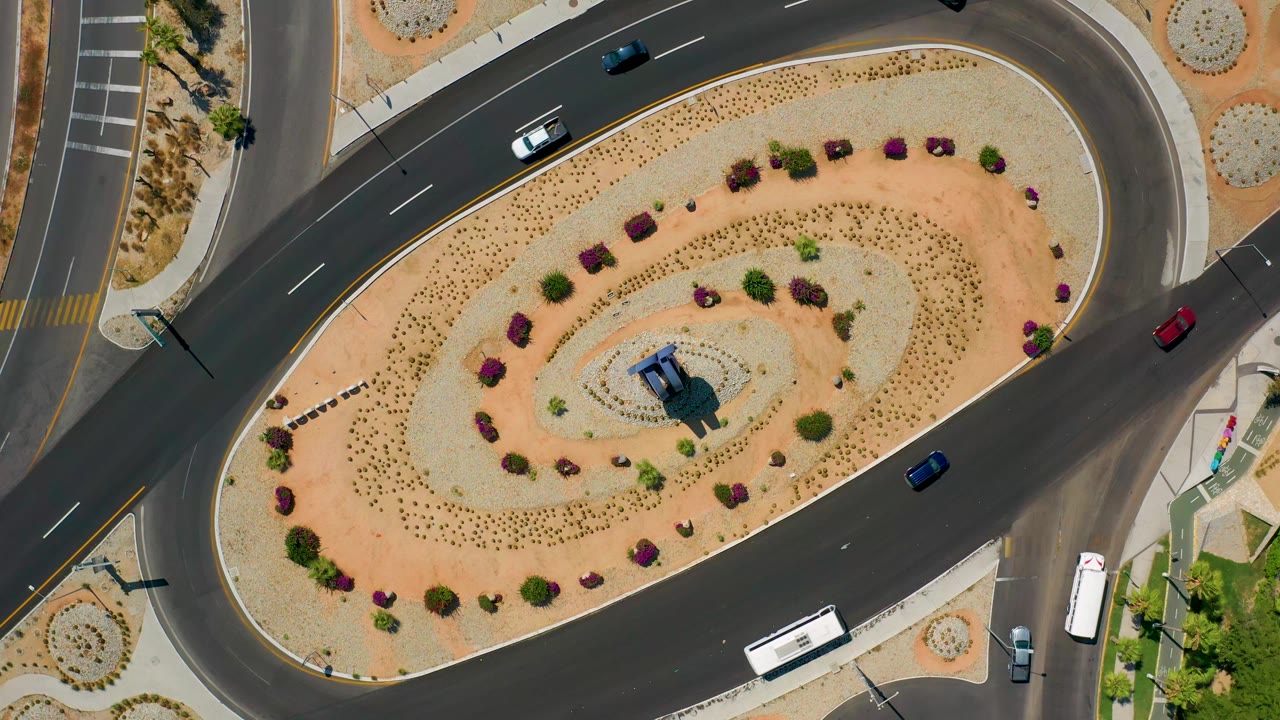 Overhead aerial frame of the Los Cabos roundabout with the landscaped center held in view, cars on the upper and right lanes, a white bus at the lower edge, and no visible people.
