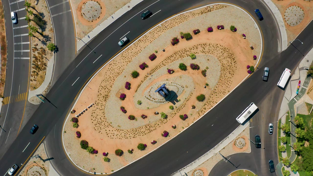 Second overhead frame in Los Cabos, Mexico, with cars spaced along the curved lanes, a white bus near the lower right road, desert landscaping, and no visible people.