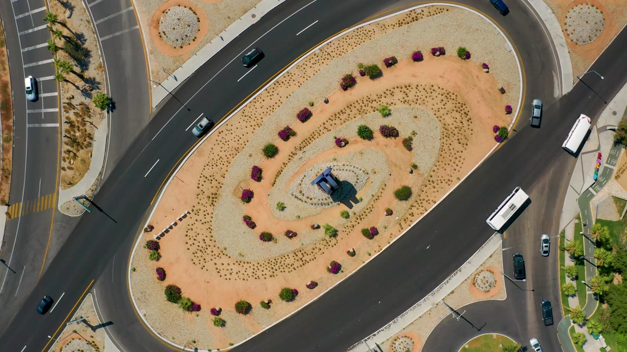 Top-down Los Cabos, Mexico frame showing the white bus higher on the right lane, multiple cars on the surrounding roads, patterned shrubs in the center island, and no visible people.