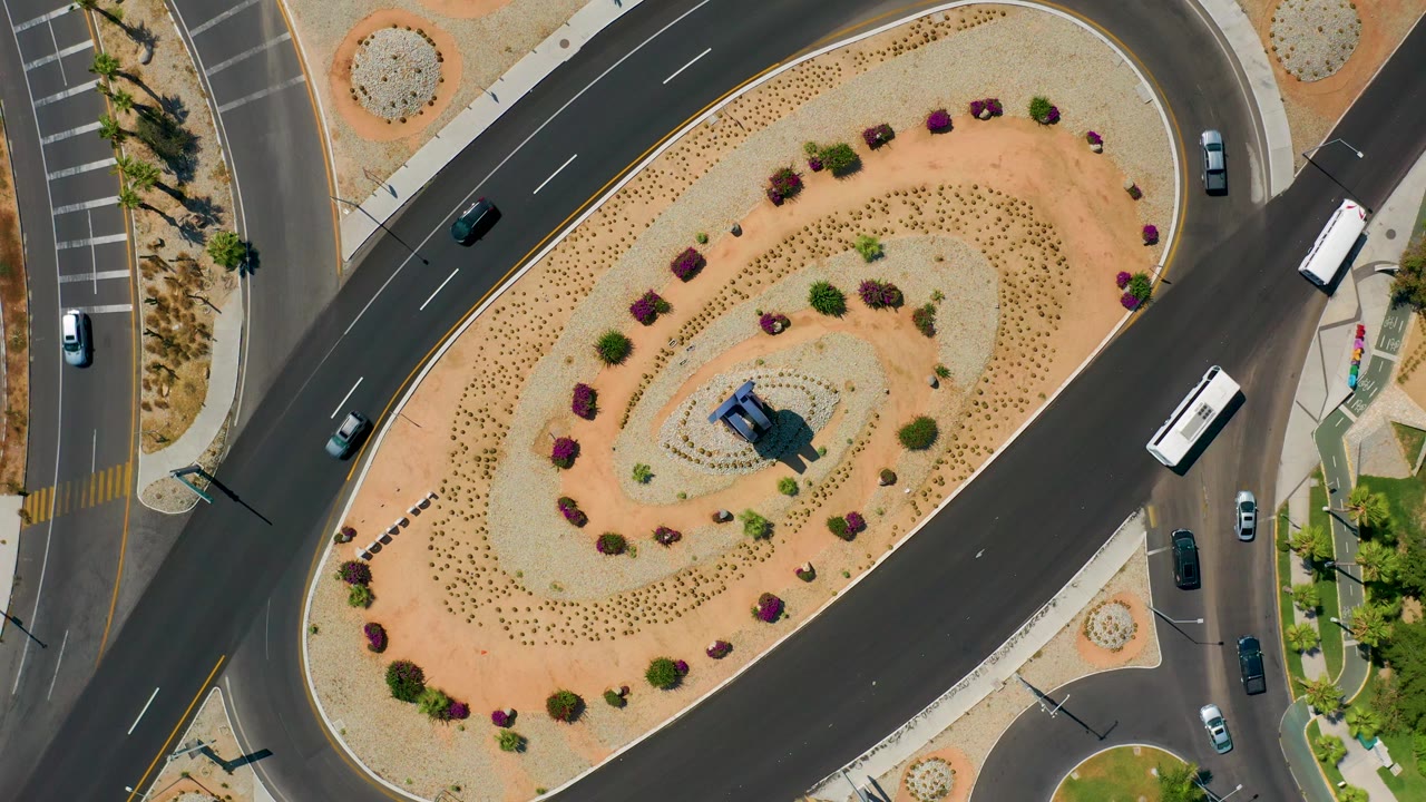 Aerial vertical frame in Los Cabos, Mexico, with the oval island centered, cars on the left and right approach roads, a white bus near the right curb, and no visible people.