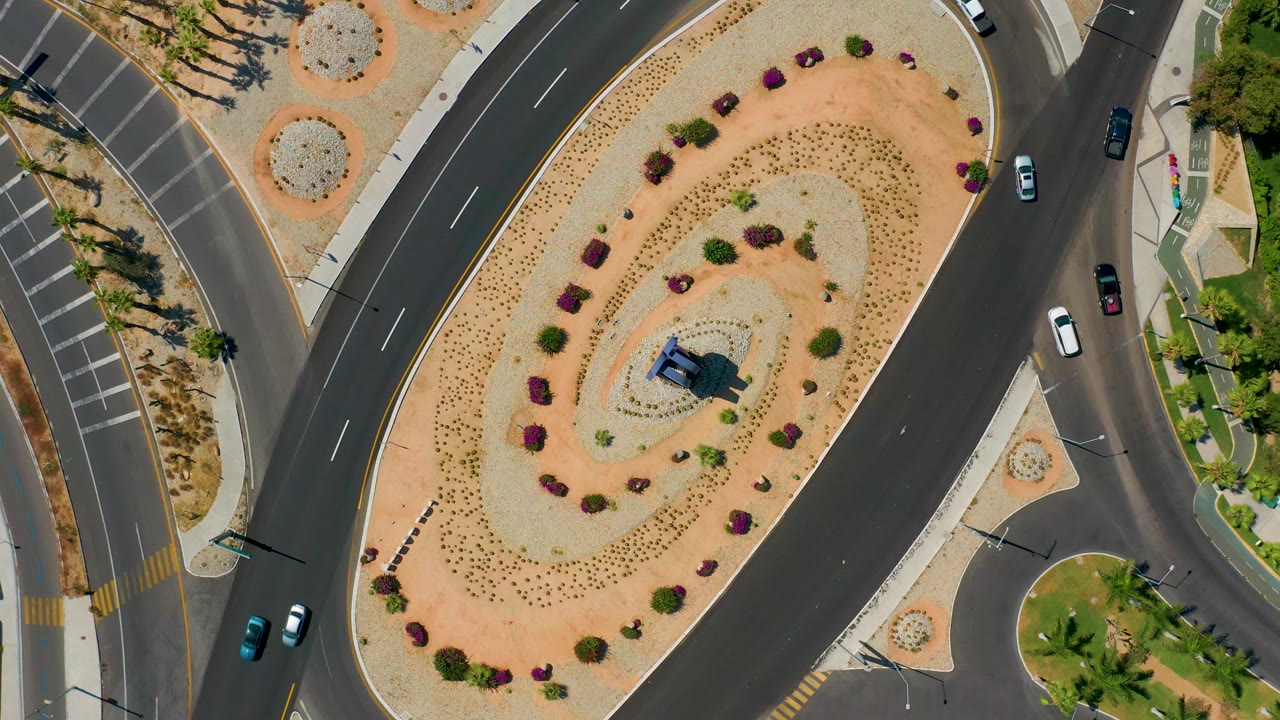 A Los Cabos aerial frame centers the oval planted island while cars appear on the surrounding lanes and no visible people are present.
