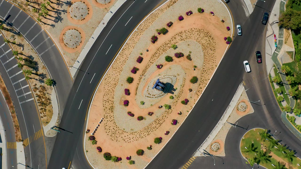 Top-down Los Cabos road view with a large landscaped roundabout, a white vehicle on the right lane, parked-space striping, palms, and no visible people.