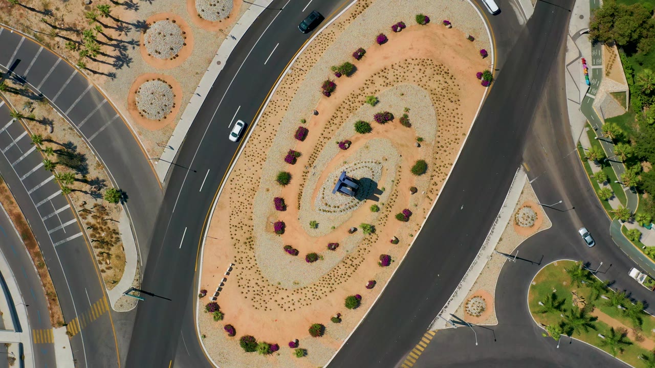Overhead Los Cabos road frame with a white car on the left curve, a dark vehicle near the top road, a landscaped oval median, parking stripes, and no visible people.