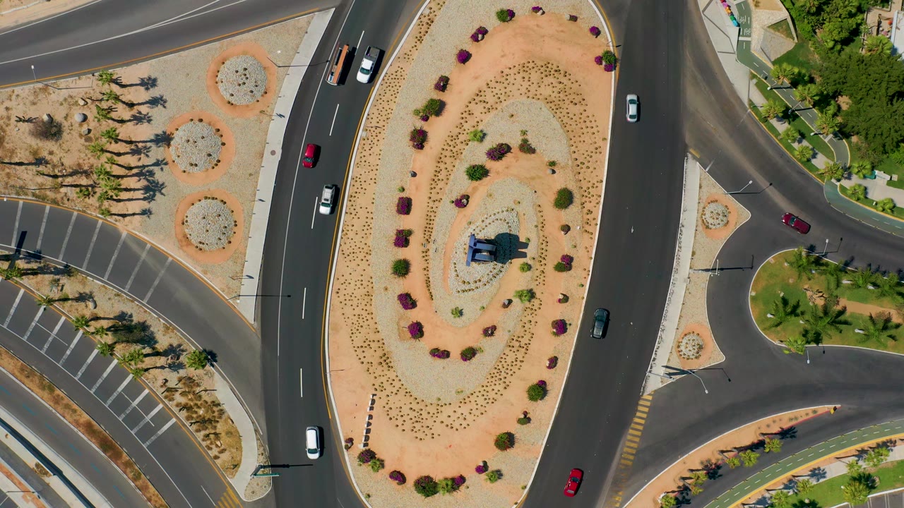 Overhead Los Cabos road view with a truck near the left lanes, cars on the curved asphalt, a spiral planting bed, and no visible people.