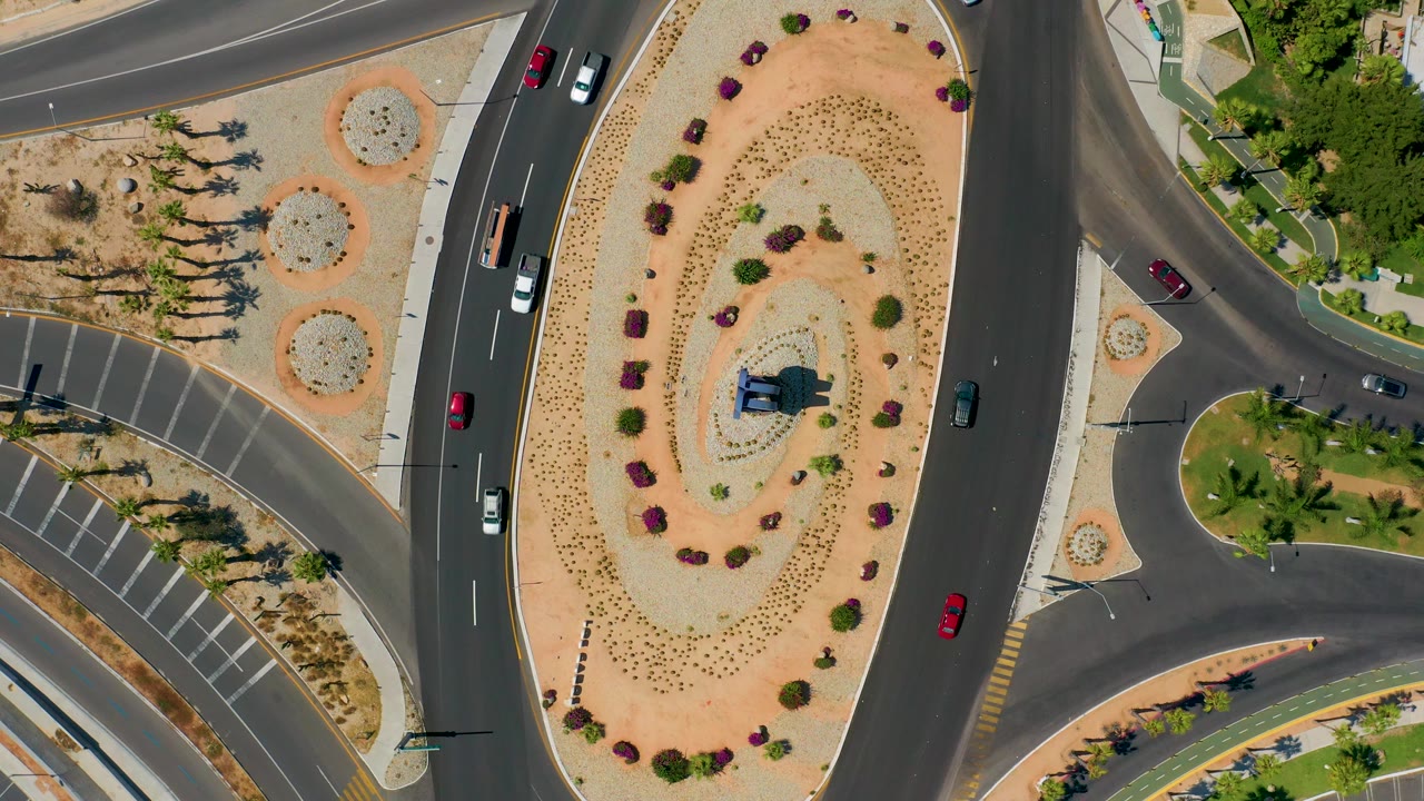 Overhead Los Cabos frame with cars on the right and upper roads, a central landscaped spiral, palm-lined edges, and no visible people.