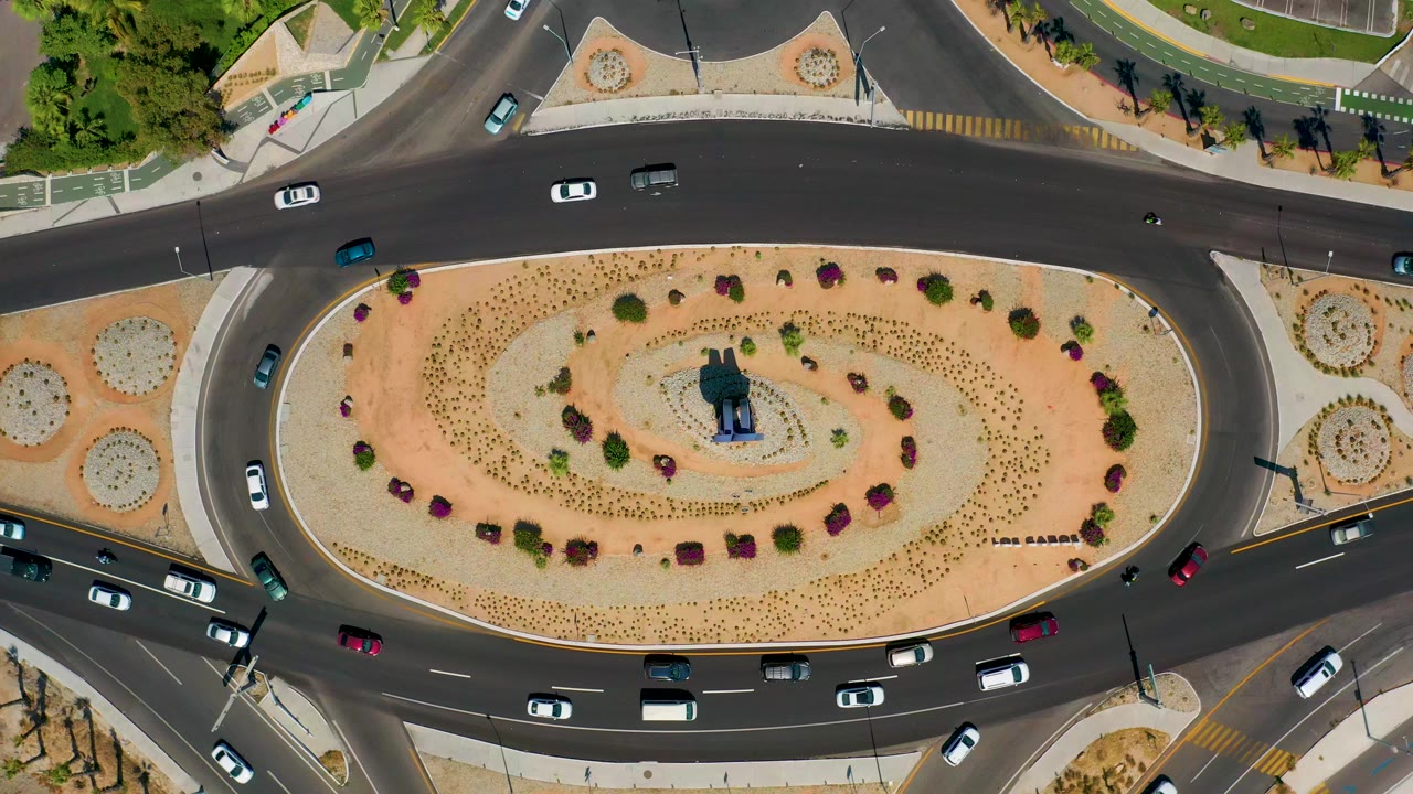 Opening overhead frame of a landscaped oval roundabout in Los Cabos, Mexico, with cars spaced around dark asphalt lanes and no visible people.