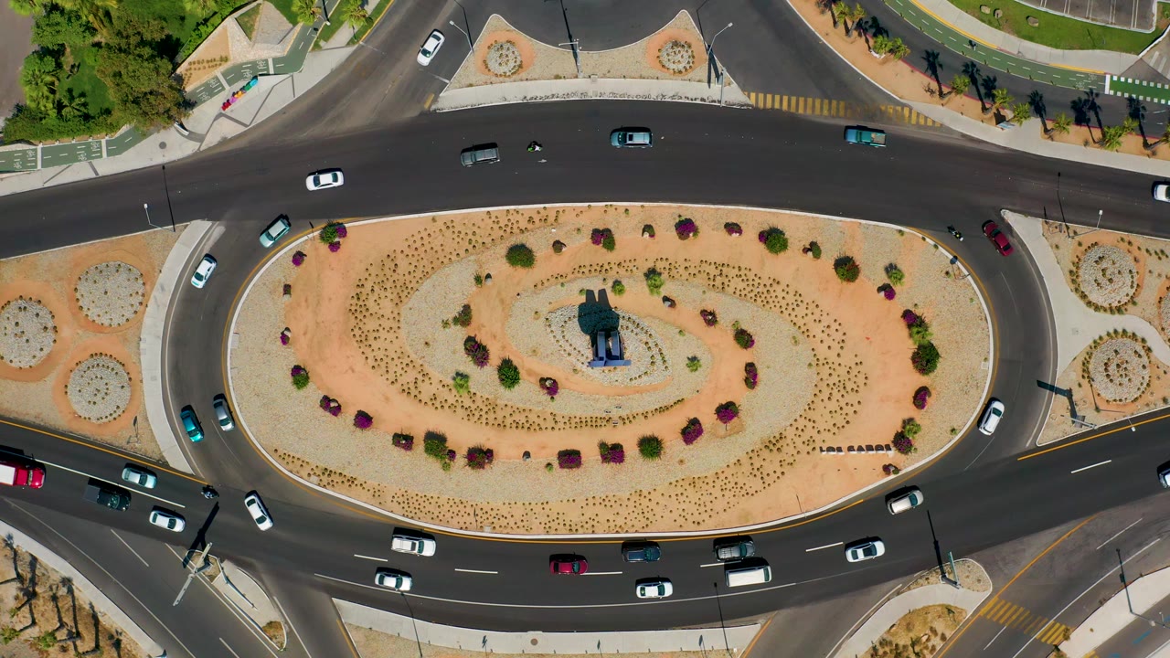A top-down Los Cabos, Mexico frame shows the oval center island, low shrubs, circular paving details, and vehicles spaced around the roundabout, with no visible people.