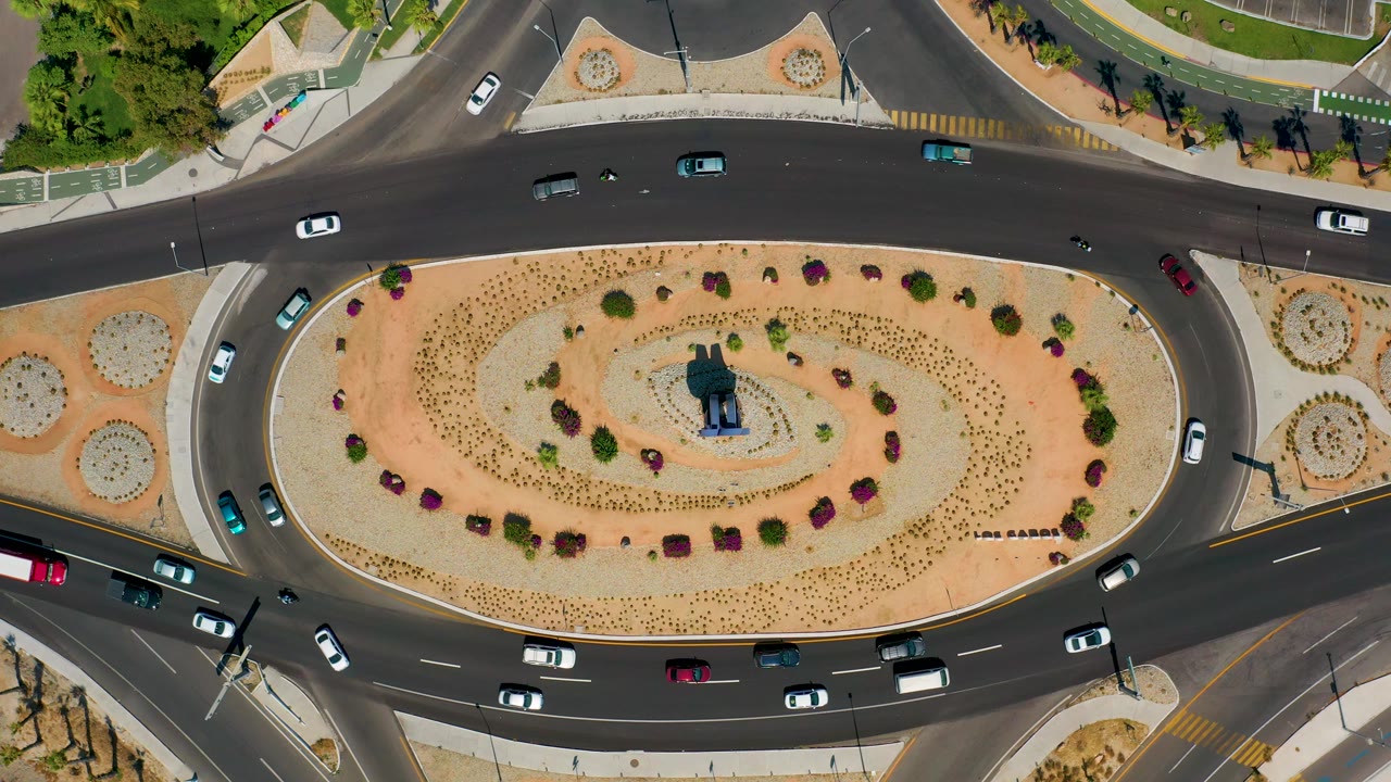 Overhead view in Los Cabos, Mexico, with cars and small motorcycles entering and exiting the roundabout beside tan landscaping, sidewalks, and painted crosswalks, with no visible people.