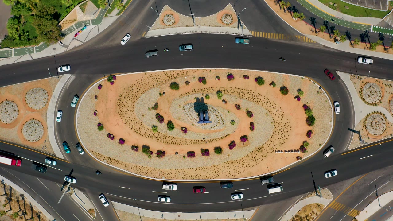 Los Cabos, Mexico overhead frame showing a landscaped traffic circle with cars on the surrounding lanes, a sandy center bed, planted shrubs, and no visible people.
