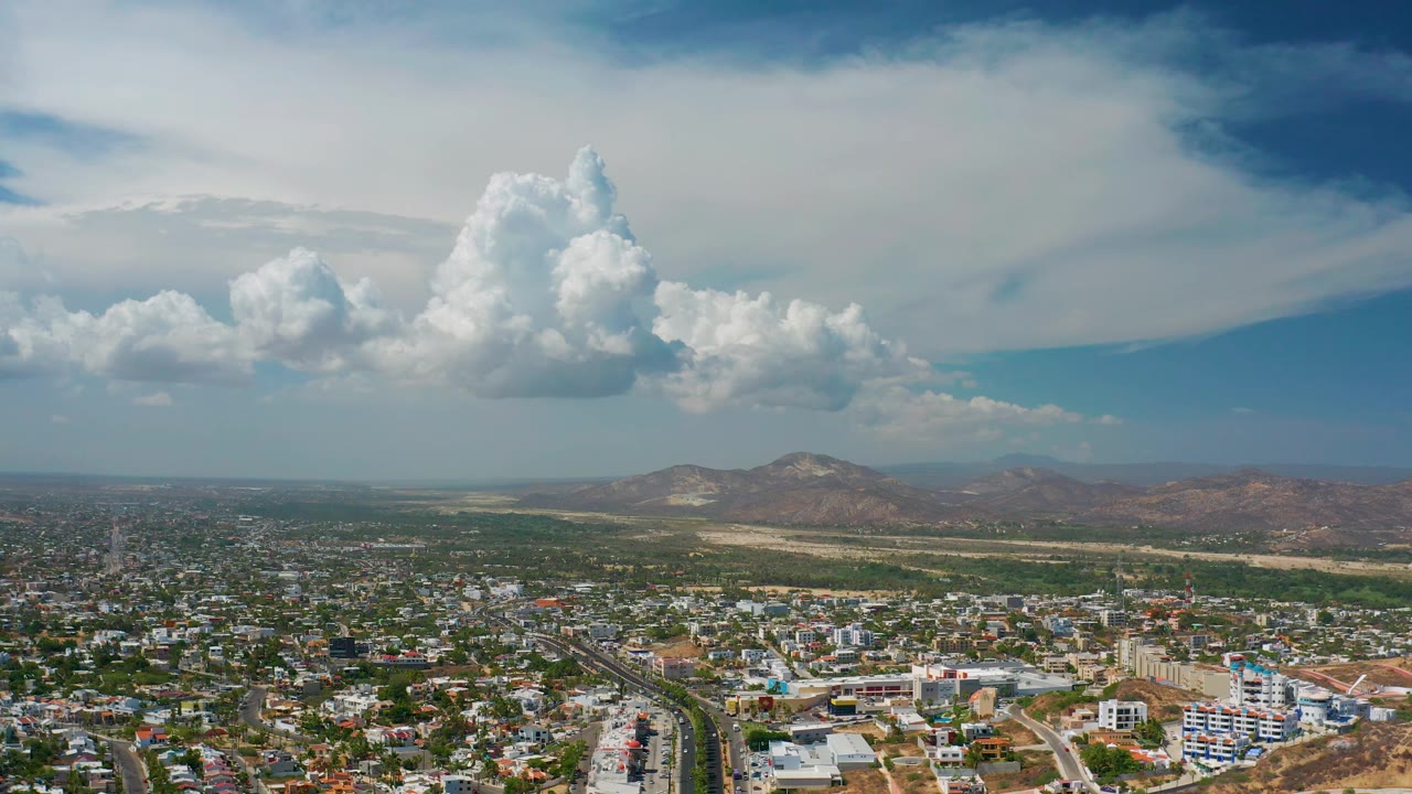 Los Cabos aerial panorama with bright low buildings in the foreground, a green strip before the hills, and high white clouds overhead, no visible people.