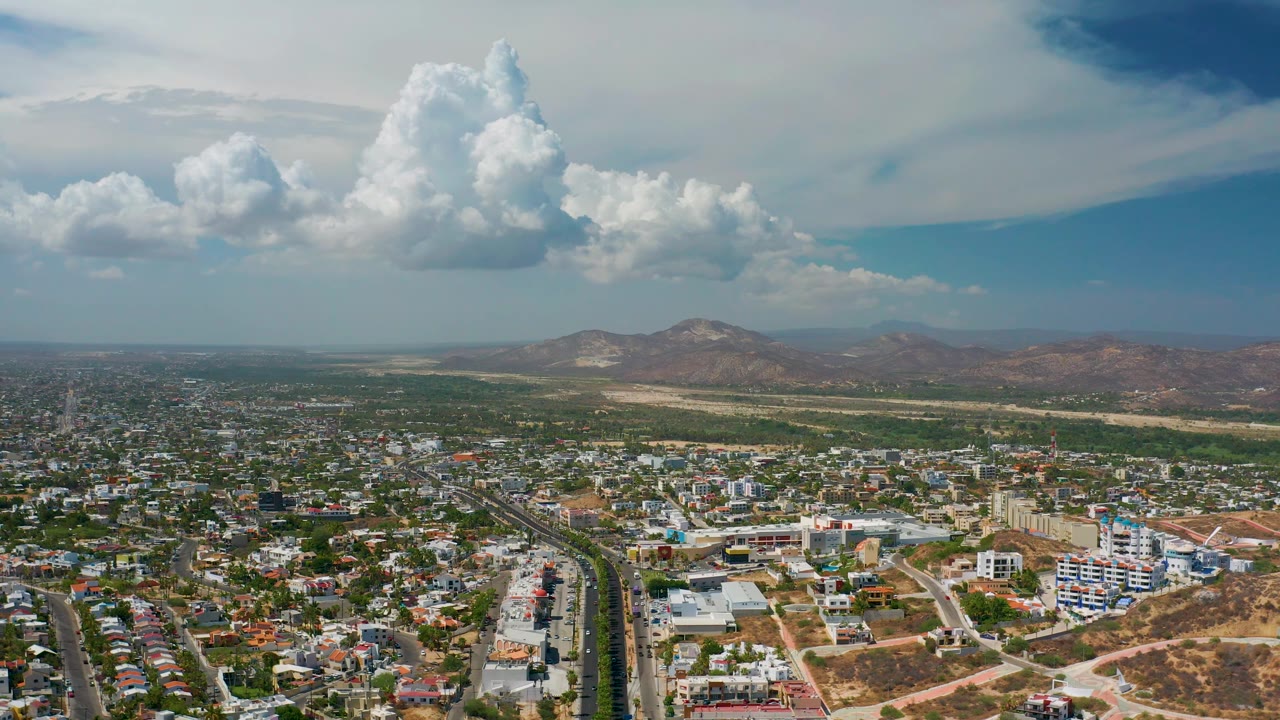 Los Cabos aerial frame with a road corridor at left, modern low-rise buildings near the center, dry hills on the horizon, layered clouds, and no visible people.
