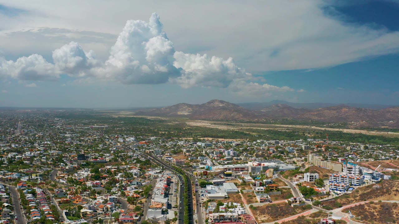 Aerial city edge with vegetation, hills, and tall clouds