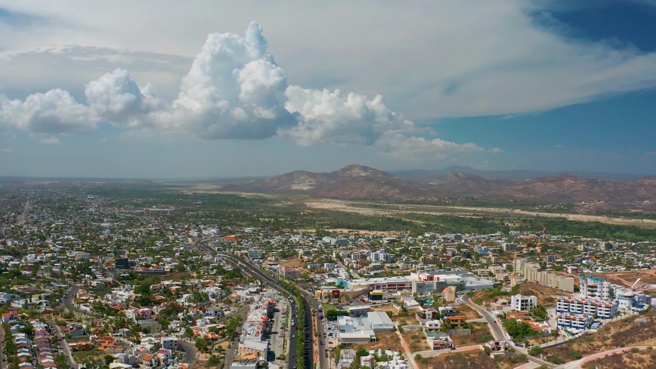 A wide aerial frame over Los Cabos, Mexico shows low-rise city blocks, a divided road, distant desert mountains, and large white clouds with no visible people.