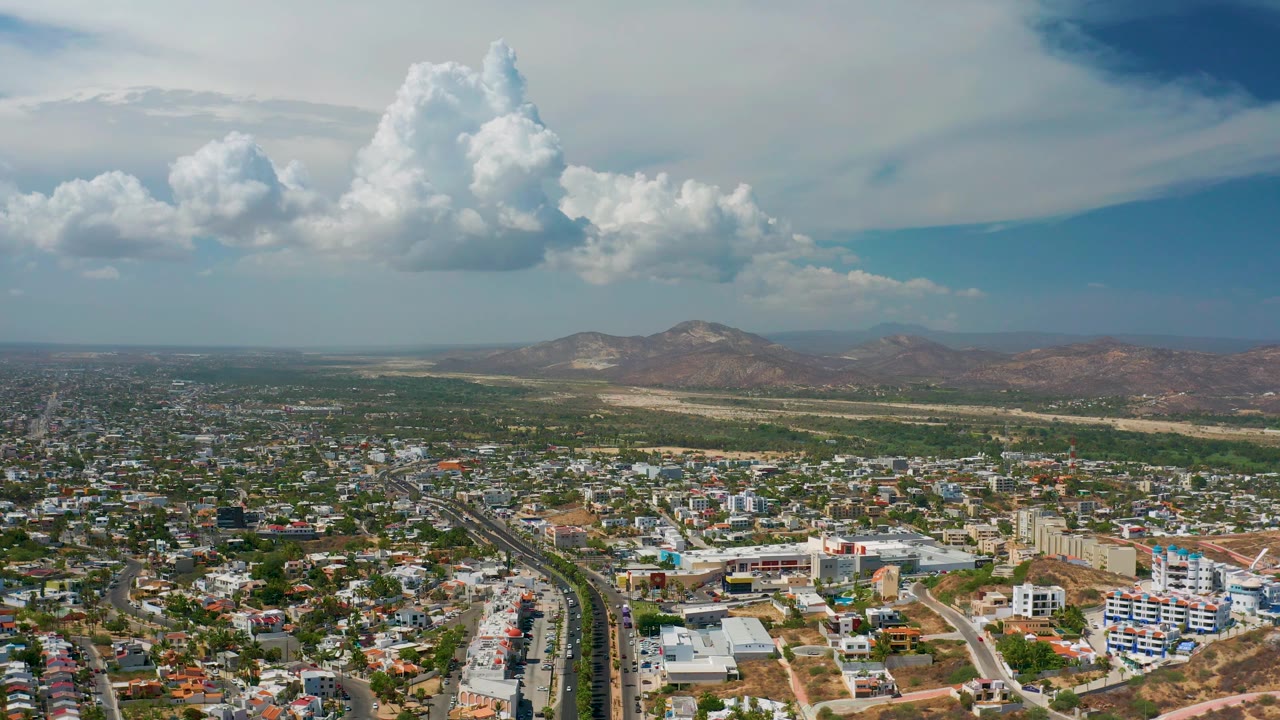 Drone-like view over city rooftops toward desert mountains under clouds