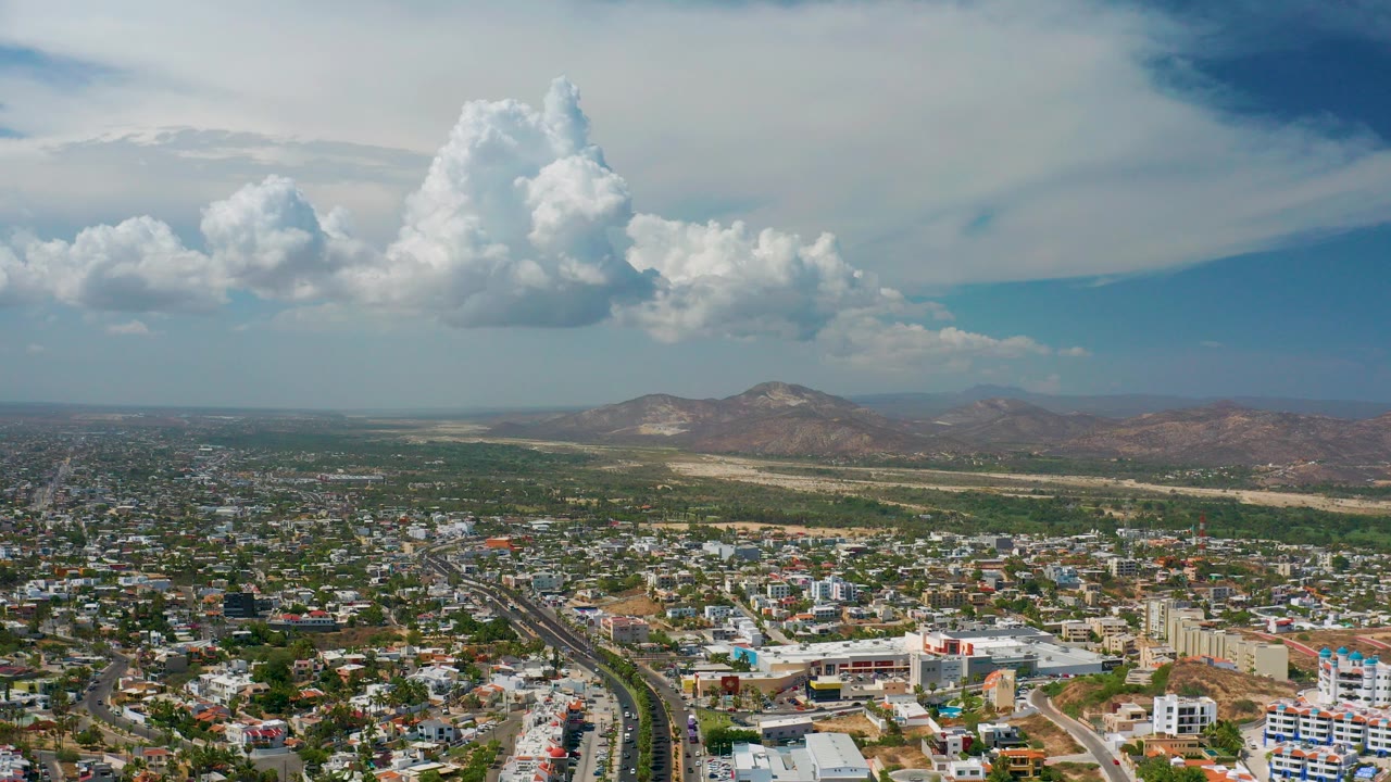 Opening aerial view over Los Cabos, Mexico, showing dense buildings and a road corridor below distant mountains and large cumulus clouds, with no visible people.