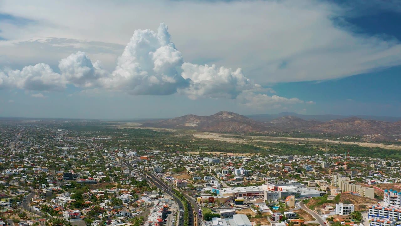 Aerial panorama of Los Cabos, Mexico, with a dark road line running through neighborhoods toward dry land, mountains, and towering clouds, with no visible people.