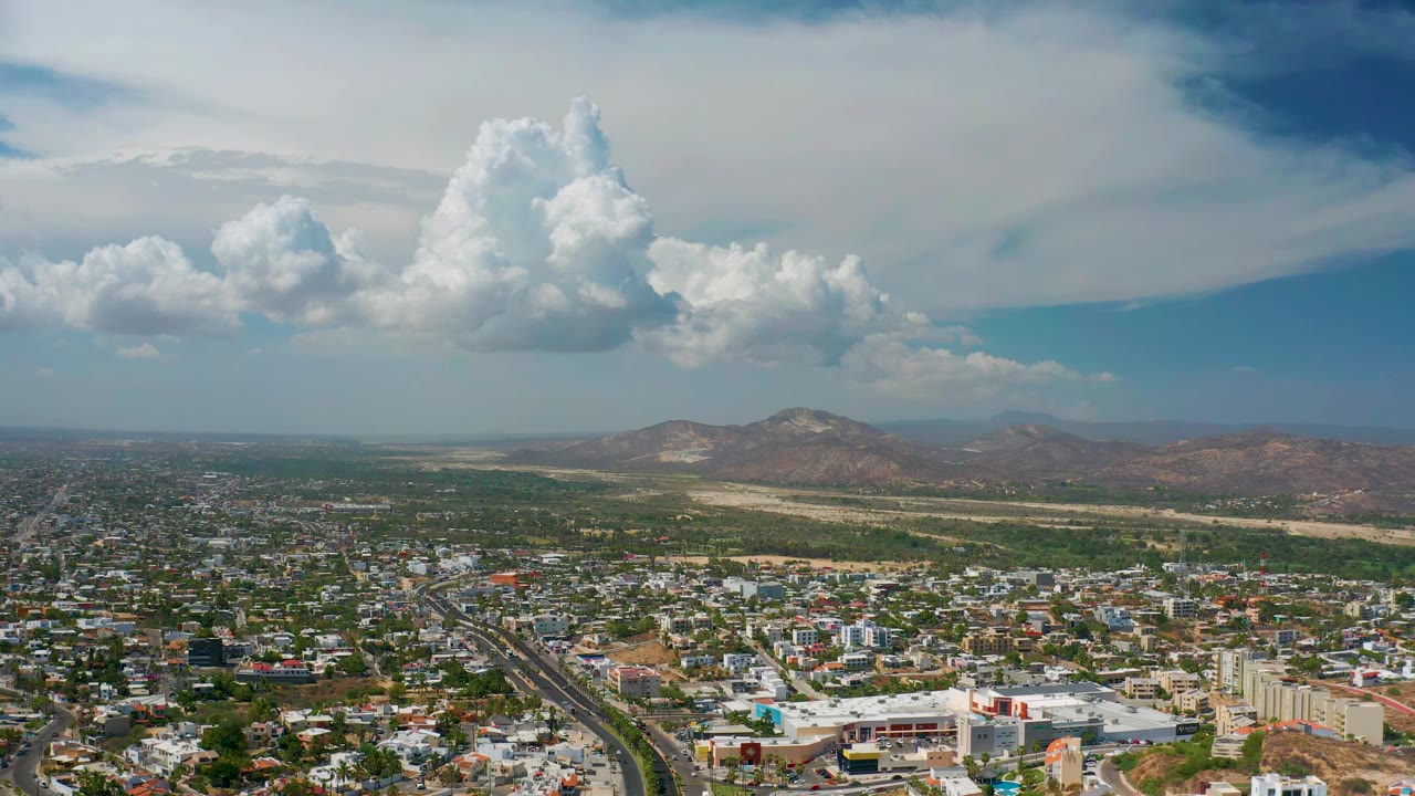 Wide aerial landscape with city blocks, hills, and towering clouds.
