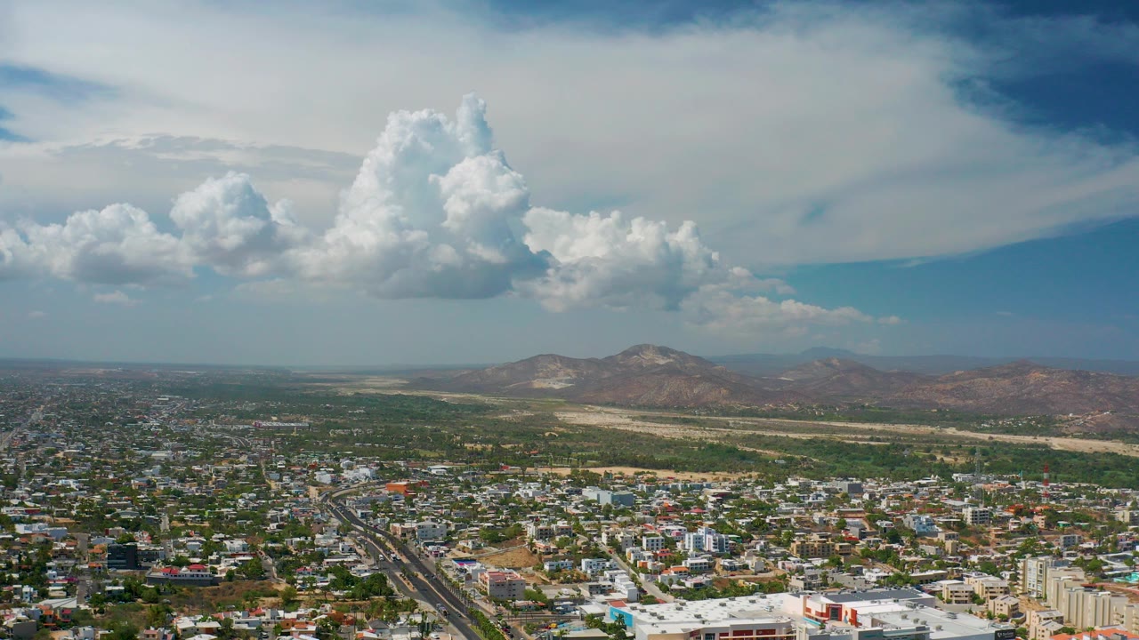Opening aerial view over Los Cabos, Mexico, with low city buildings, a road corridor, distant brown mountains, large white clouds, and no visible people.