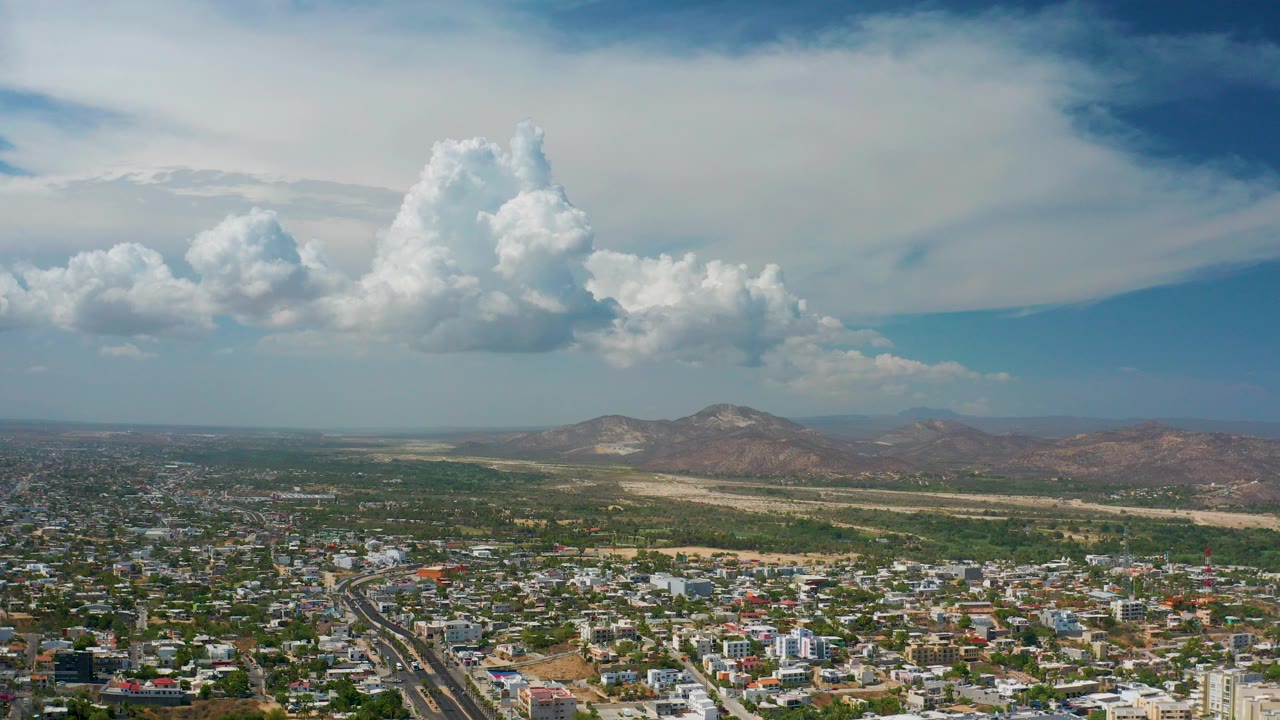 Wide aerial view over Los Cabos, Mexico, with city blocks in the foreground, dry hills and mountains on the horizon, tall white clouds overhead, and no visible people.
