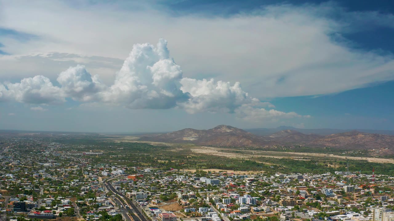 Aerial view in Los Cabos showing an urban edge, patches of vegetation, dry terrain, reddish hills, distant mountains, high cloud cover, and no visible people.