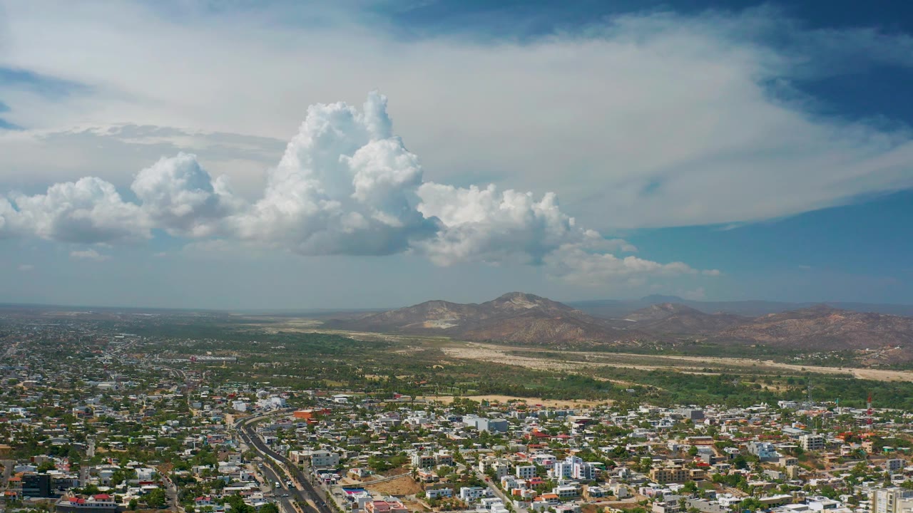 Los Cabos city foreground with a road corridor at lower left, low buildings across the frame, a mountain ridge beyond, bright clouds, and no visible people.