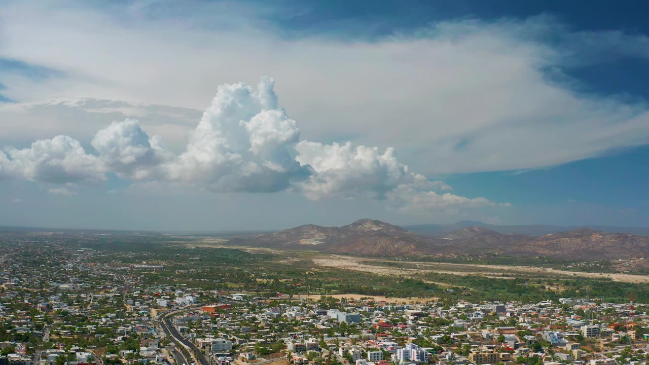 Aerial frame from Los Cabos showing dense low-rise buildings below, a pale strip of land before brown hills, and a tall central cloud with no visible people.