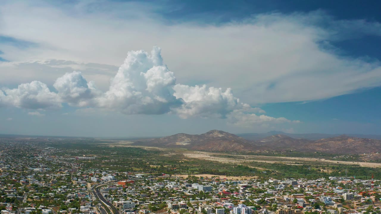 Wide Los Cabos aerial with urban blocks at the bottom edge, mountains across the background, and layered white cloud cover stretching across the sky with no visible people.