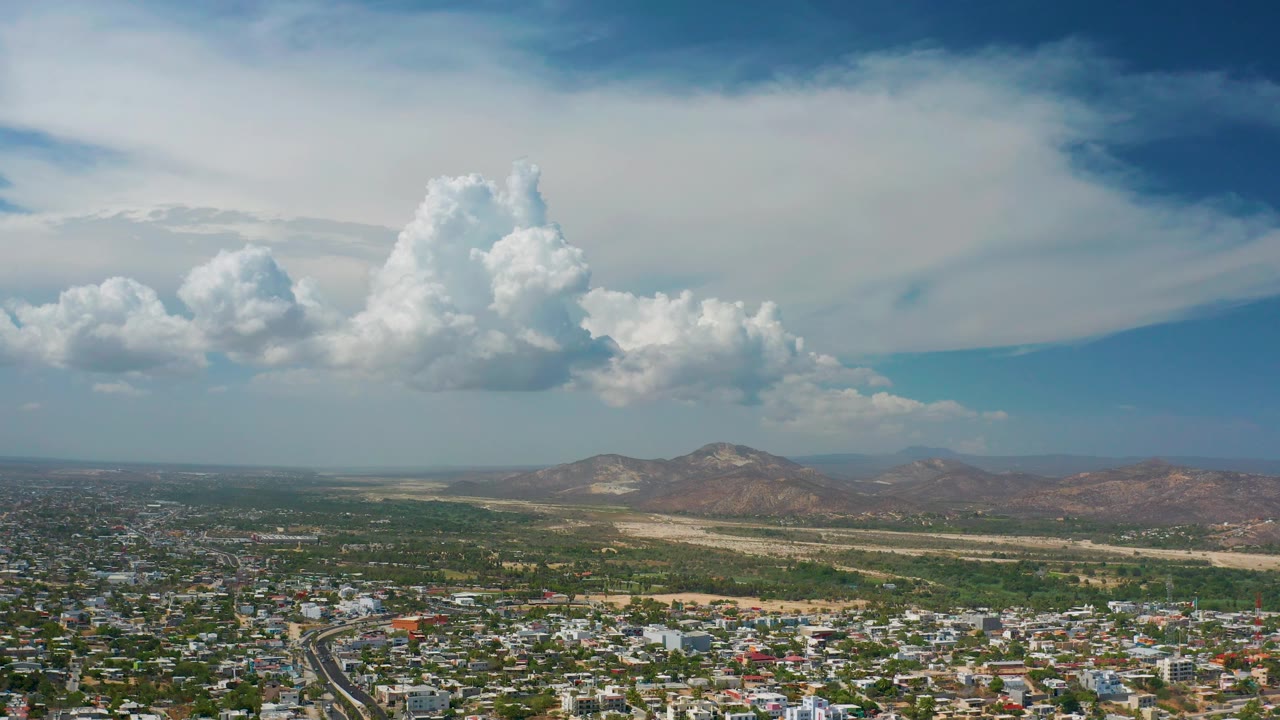Mid-clip aerial view of Los Cabos with city blocks and roads in the foreground, green terrain beyond, low hills in back, and no visible people.