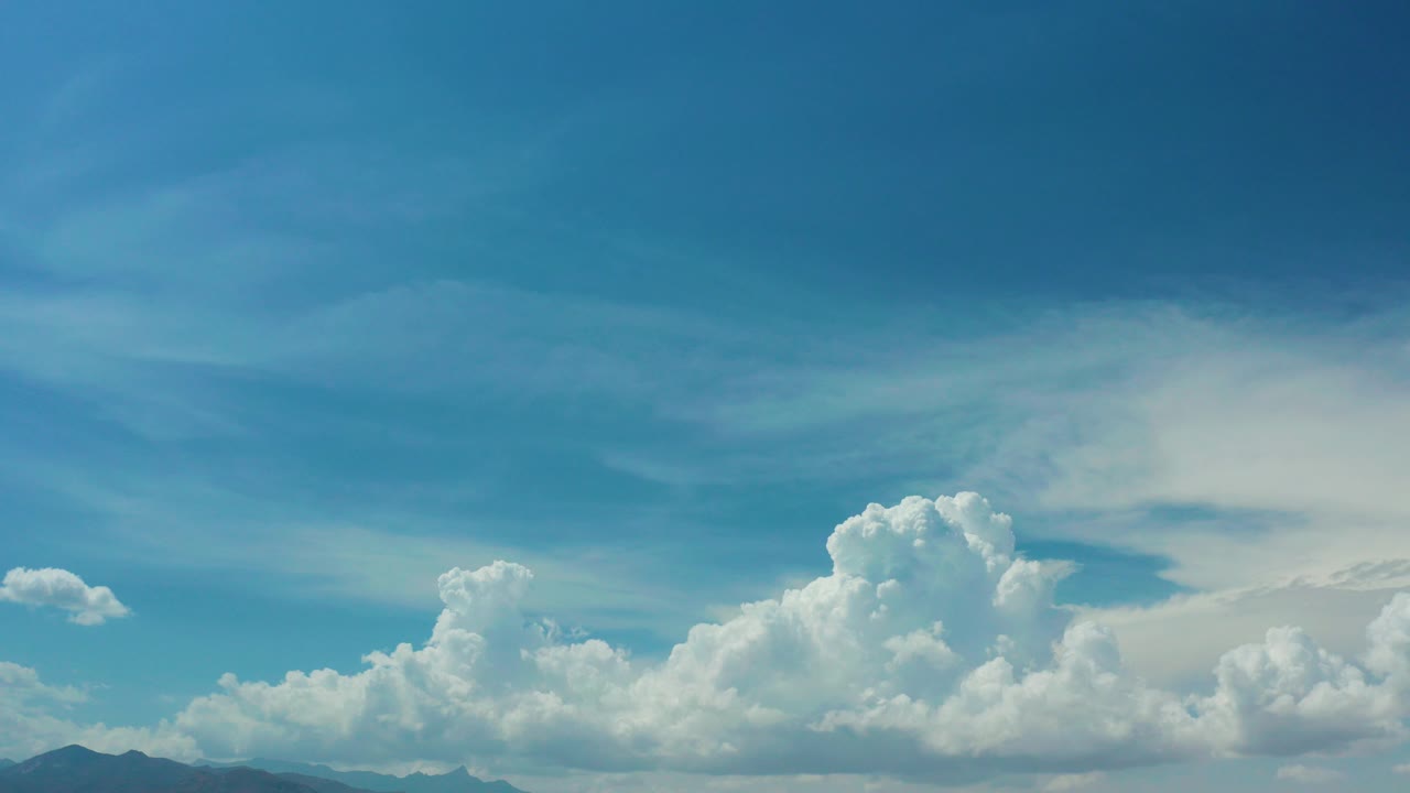Los Cabos, Mexico view of bright cumulus clouds across a blue sky with distant mountain tops low in frame and no visible people.