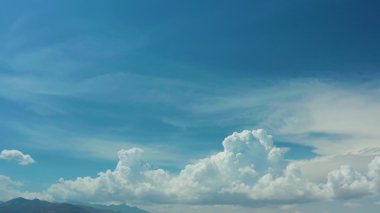Los Cabos, Mexico frame showing a wide partly cloudy sky, bright cloud clusters near the horizon, dark mountain tops at the lower left, and no visible people.