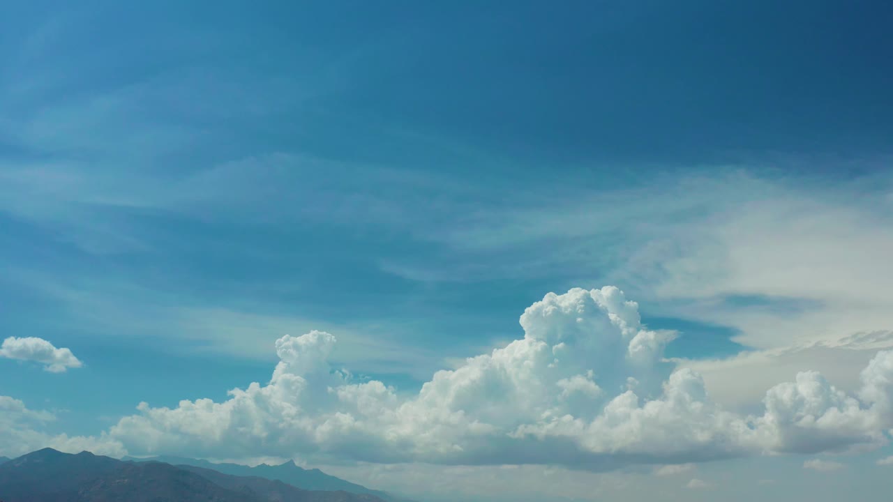 Los Cabos cloudscape with a broad cumulus bank, teal-blue sky, hazy mountain silhouettes, and no visible people one second in.
