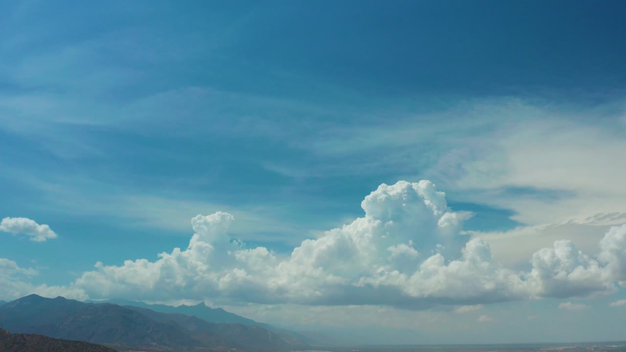 Wide Los Cabos, Mexico aerial view with a deep blue sky, tall white cloud bank, hazy mountains, distant lowlands, and no visible people.