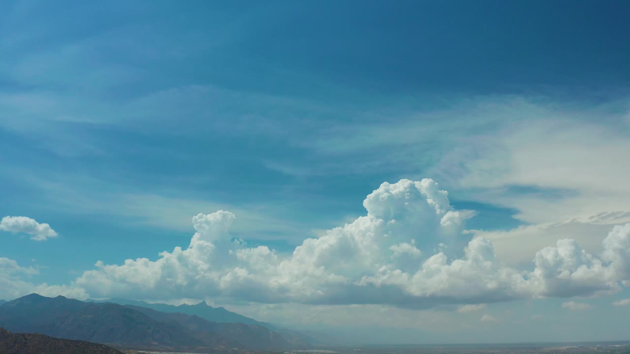 A wide Los Cabos, Mexico view shows bright cumulus clouds stretching across the sky above blue-gray mountains and low coastal terrain, with no visible people.