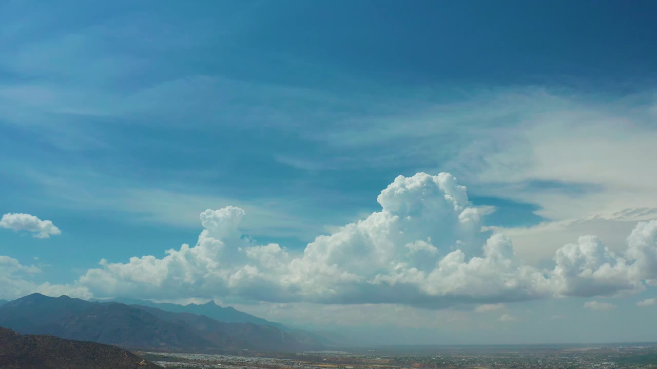Los Cabos, Mexico view with a broad blue sky, layered white clouds, dark mountain silhouettes, and no visible people.