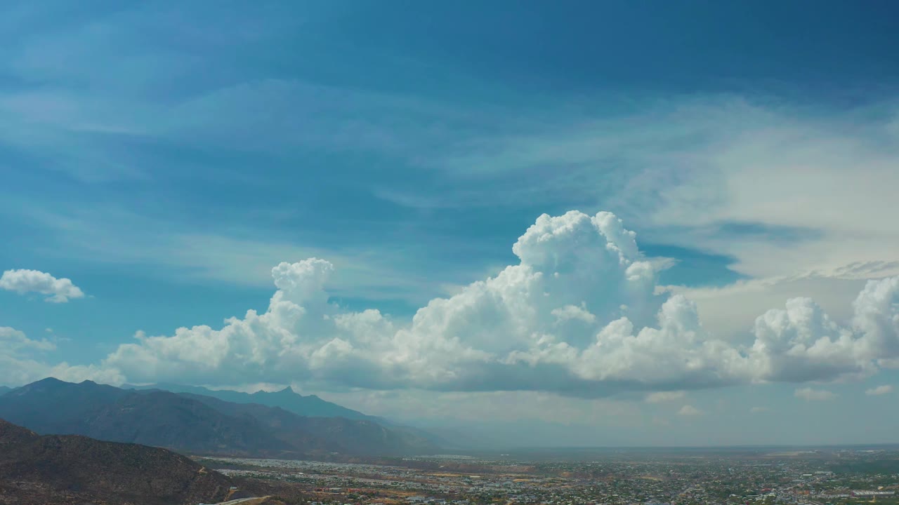Los Cabos aerial view with the cloud bank centered higher in the sky, dark mountain ridges on the left, haze near the horizon, and no visible people.