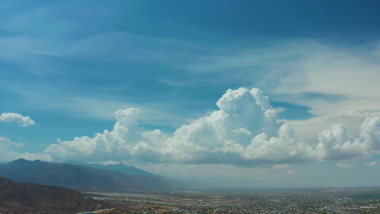 Wide Los Cabos landscape frame with bright cumulus clouds stretching across the upper right, layered mountains to the left, urban grid below, and no visible people.