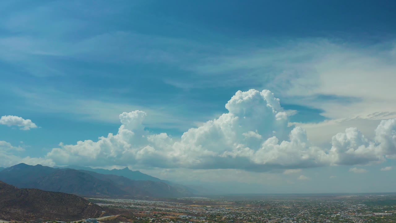 Mid-clip view of Los Cabos with soft blue sky, a dense white cloud formation, mountain slopes at left, low buildings across the valley, and no visible people.