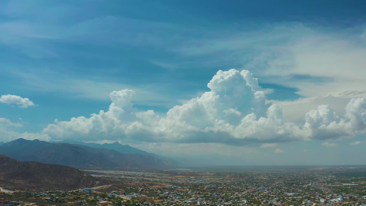 Second frame of the Los Cabos aerial view, with the built-up valley stretching across the bottom, mountain ridges on the left, a hazy horizon, large cumulus clouds, and no visible people.