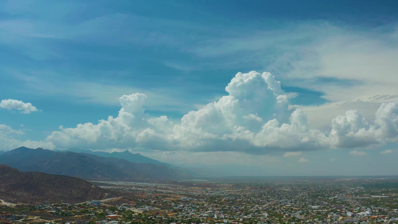 Third frame shows Los Cabos, Mexico from a low aerial angle, with city blocks across the foreground, a dark hillside at left, distant mountain silhouettes, bright clouds, and no visible people.